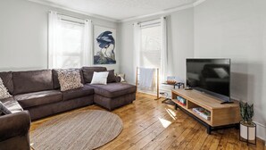 Un soggiorno moderno con un divano componibile, una TV a schermo piatto, parquet e ampie finestre con persiane.