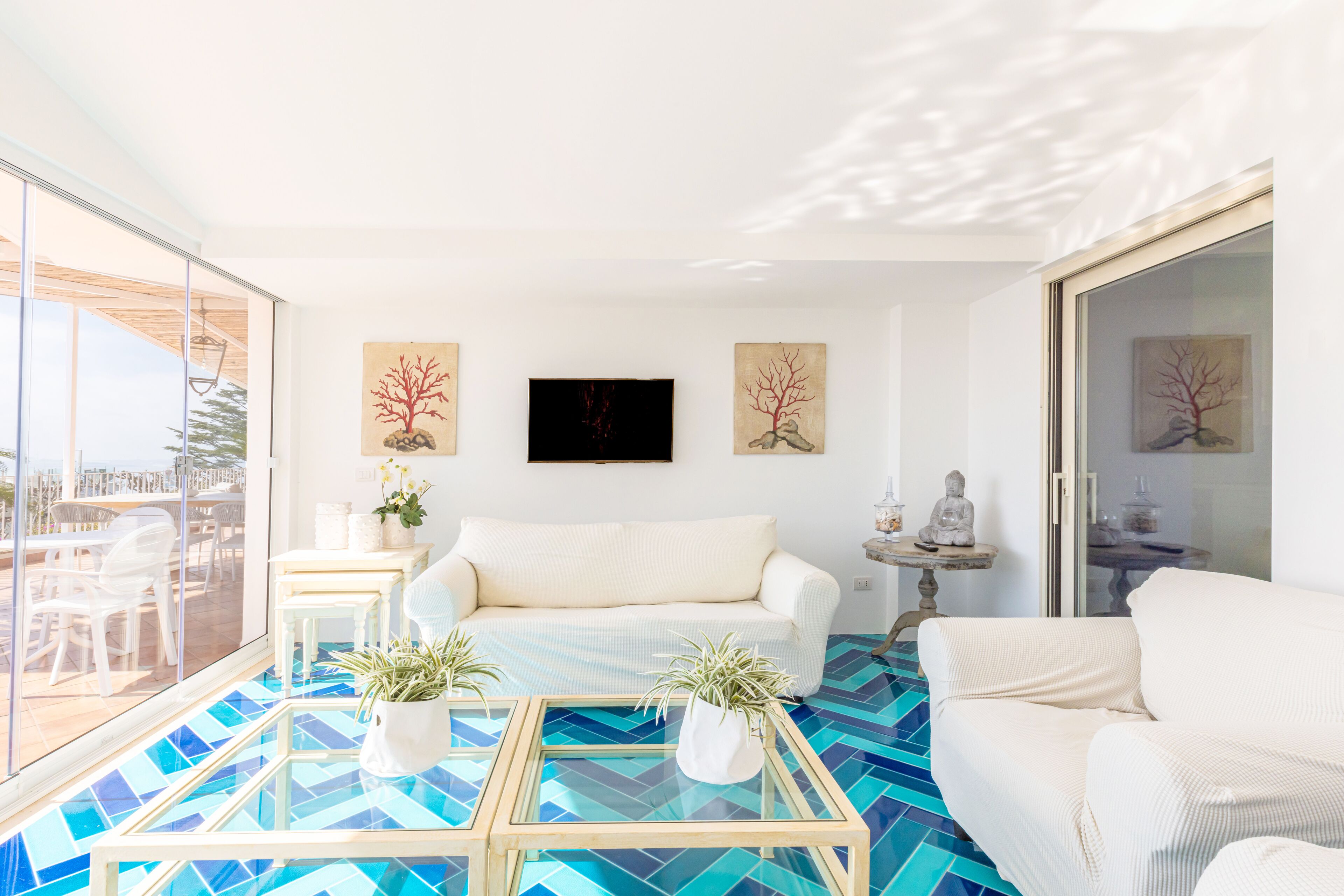 Un salon moderne avec un canapé blanc, une table basse en verre et des plantes en pot. Des tableaux ornent le mur et un téléviseur est fixé au-dessus du canapé.