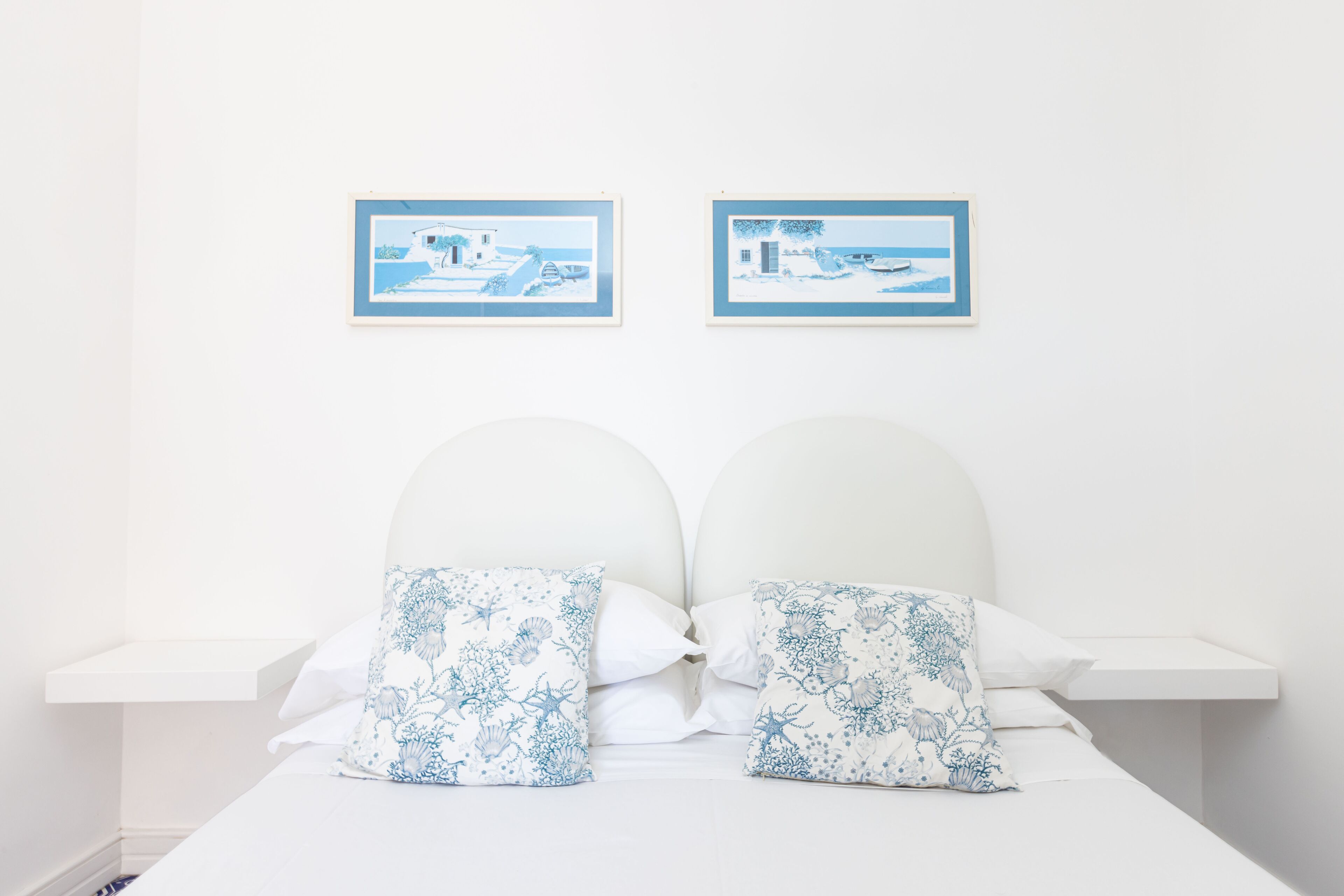 A bedroom with a white bed, two pillows with a blue floral pattern, and two framed pictures on the wall.
