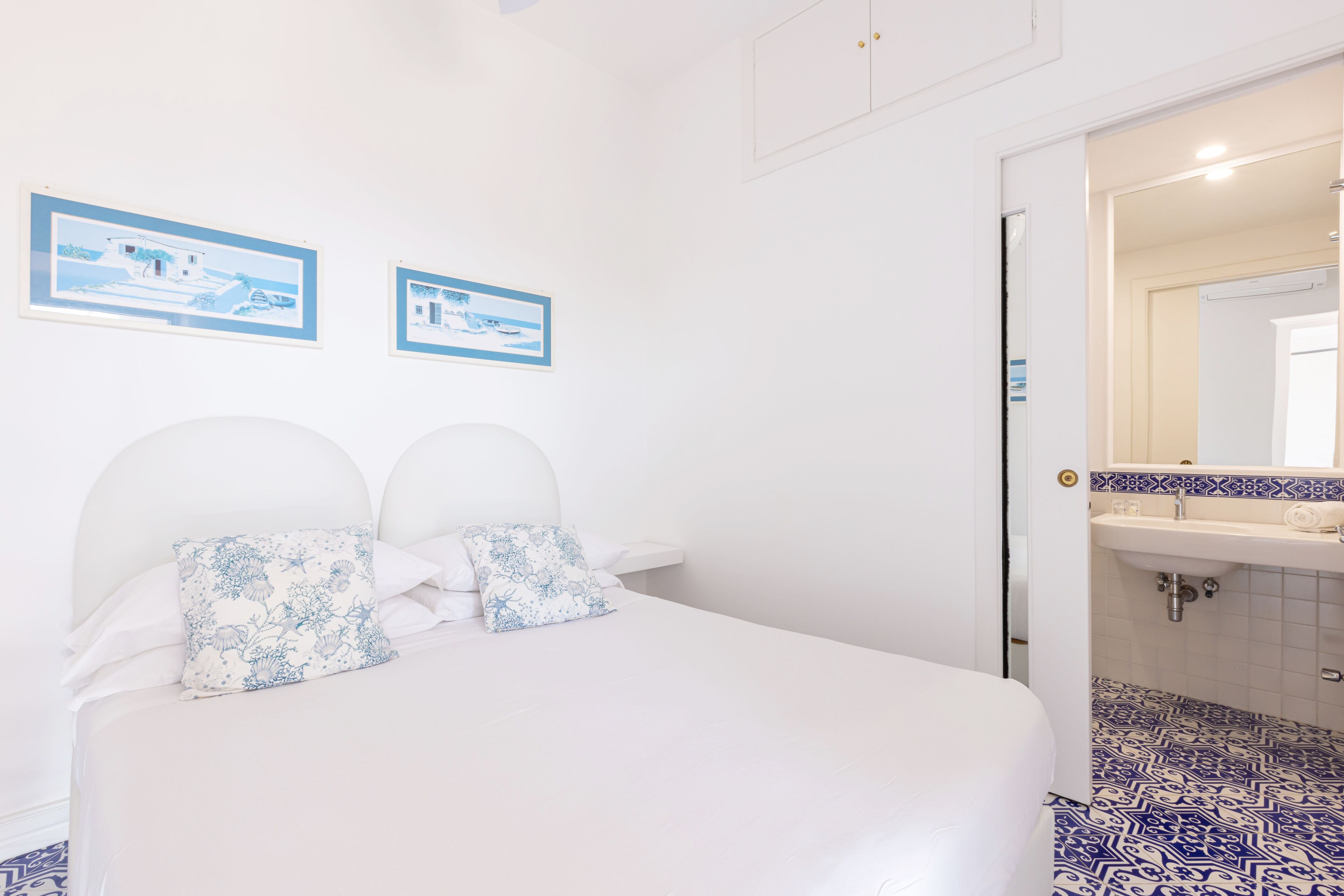 Une chambre à coucher comprenant un lit, deux oreillers, une table de chevet, un tableau accroché au mur, et une salle de bain avec un lavabo et un sol carrelé.