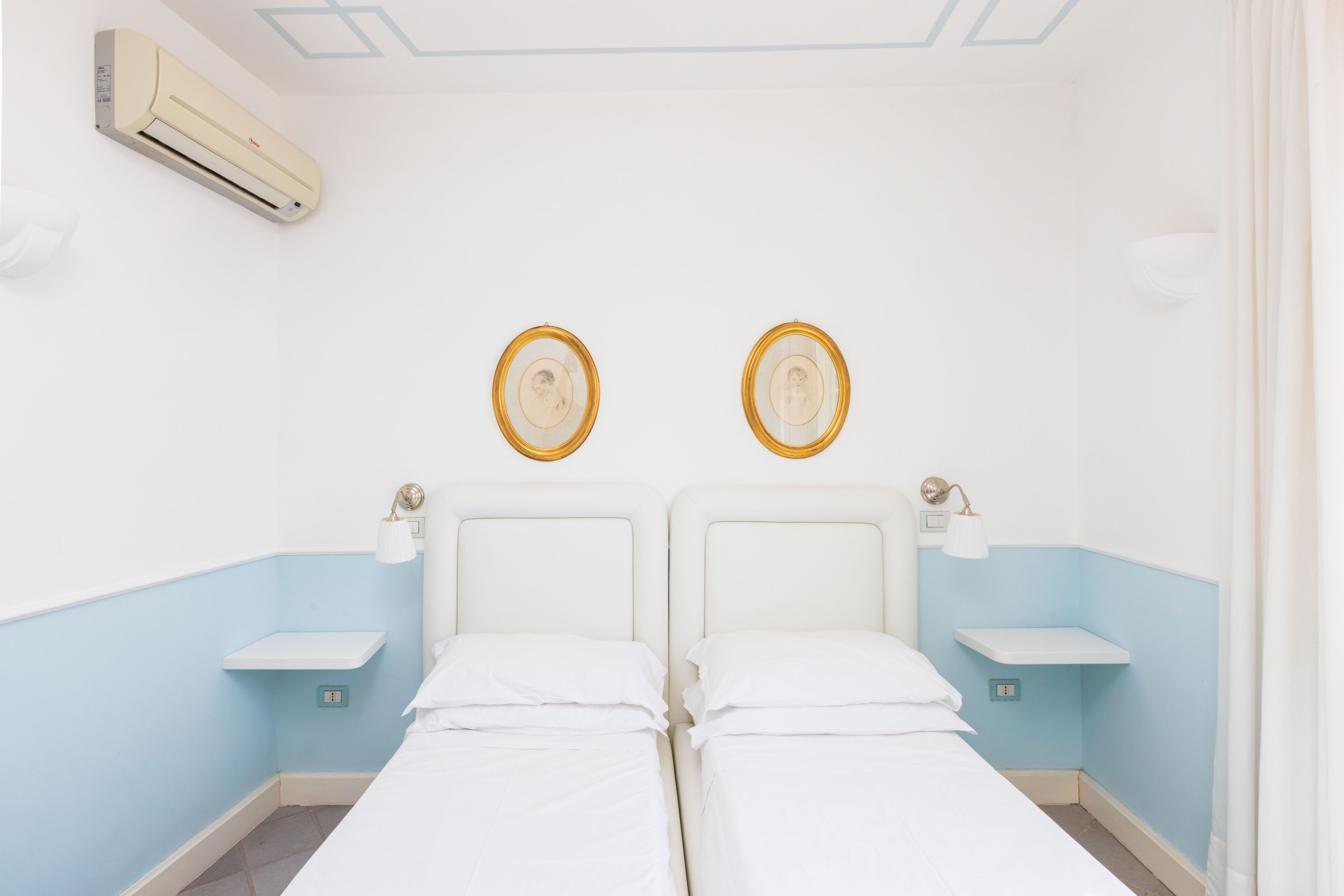 Une chambre d’hôtel avec deux lits, un climatiseur fixé au mur, des tables de chevet avec des lampes et un interrupteur sur le mur.