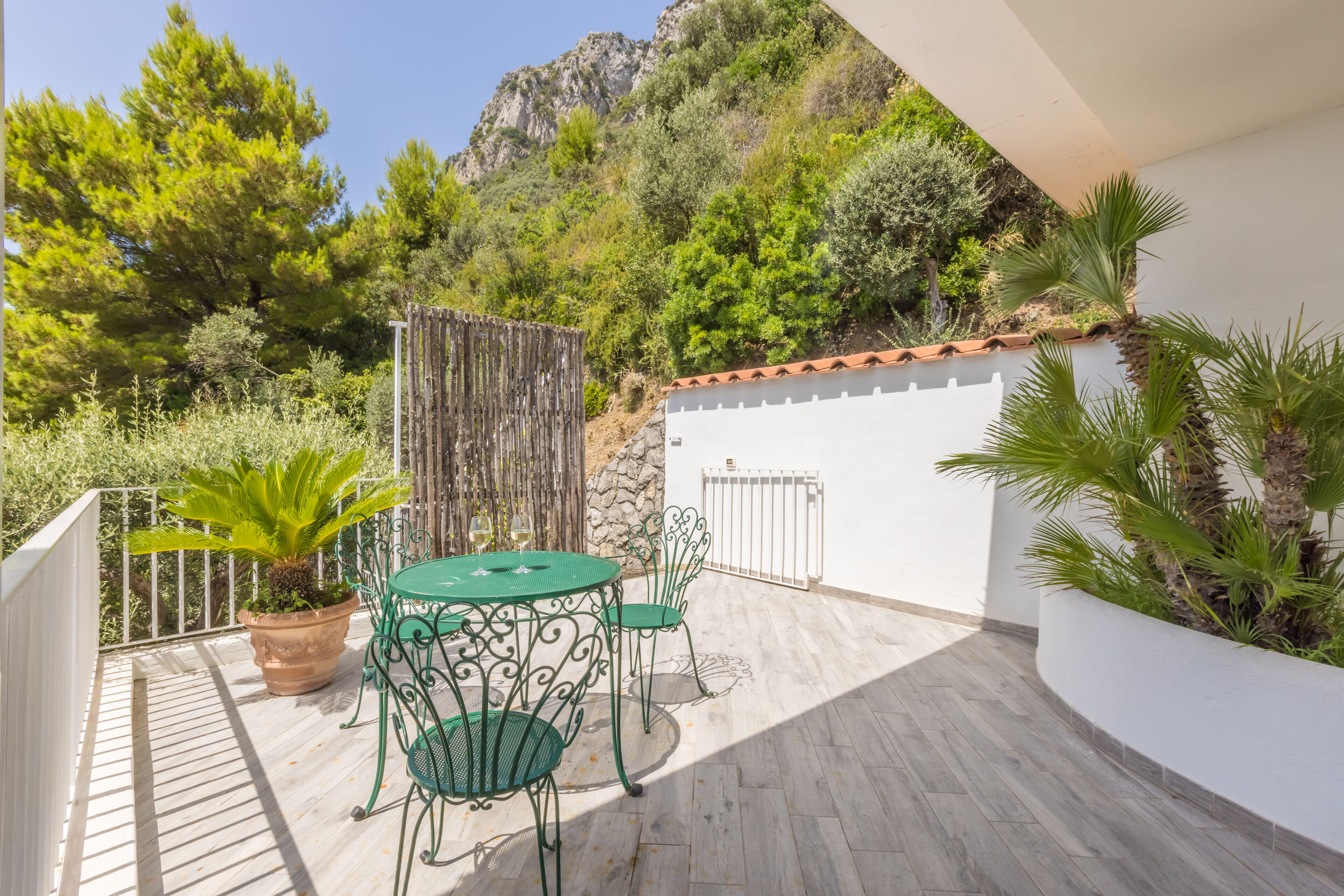 An outdoor patio with a round table and chairs, surrounded by potted plants and a white wall.