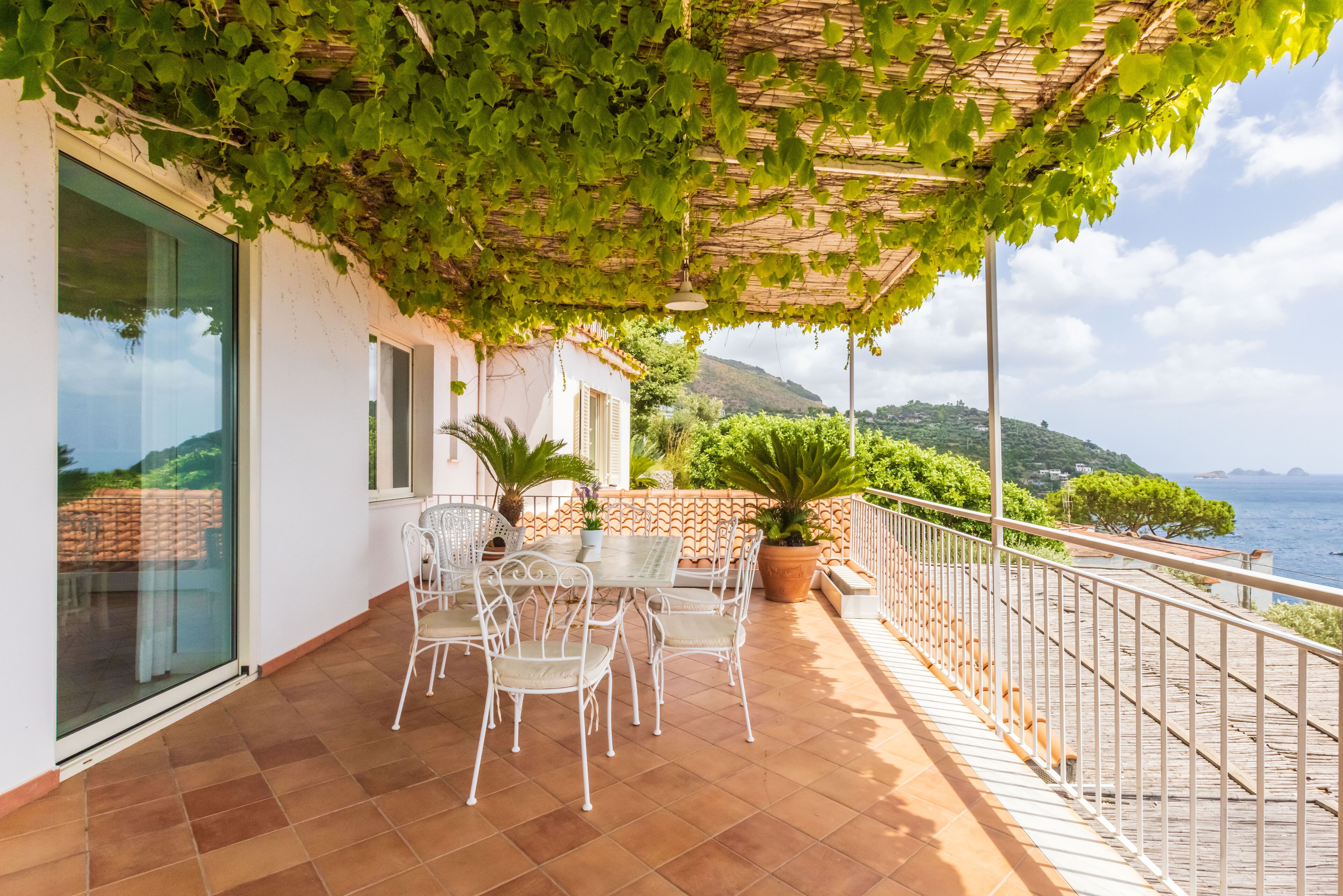 A terrace with a table and chairs, a pergola with greenery, and a view of the sea and hills.
