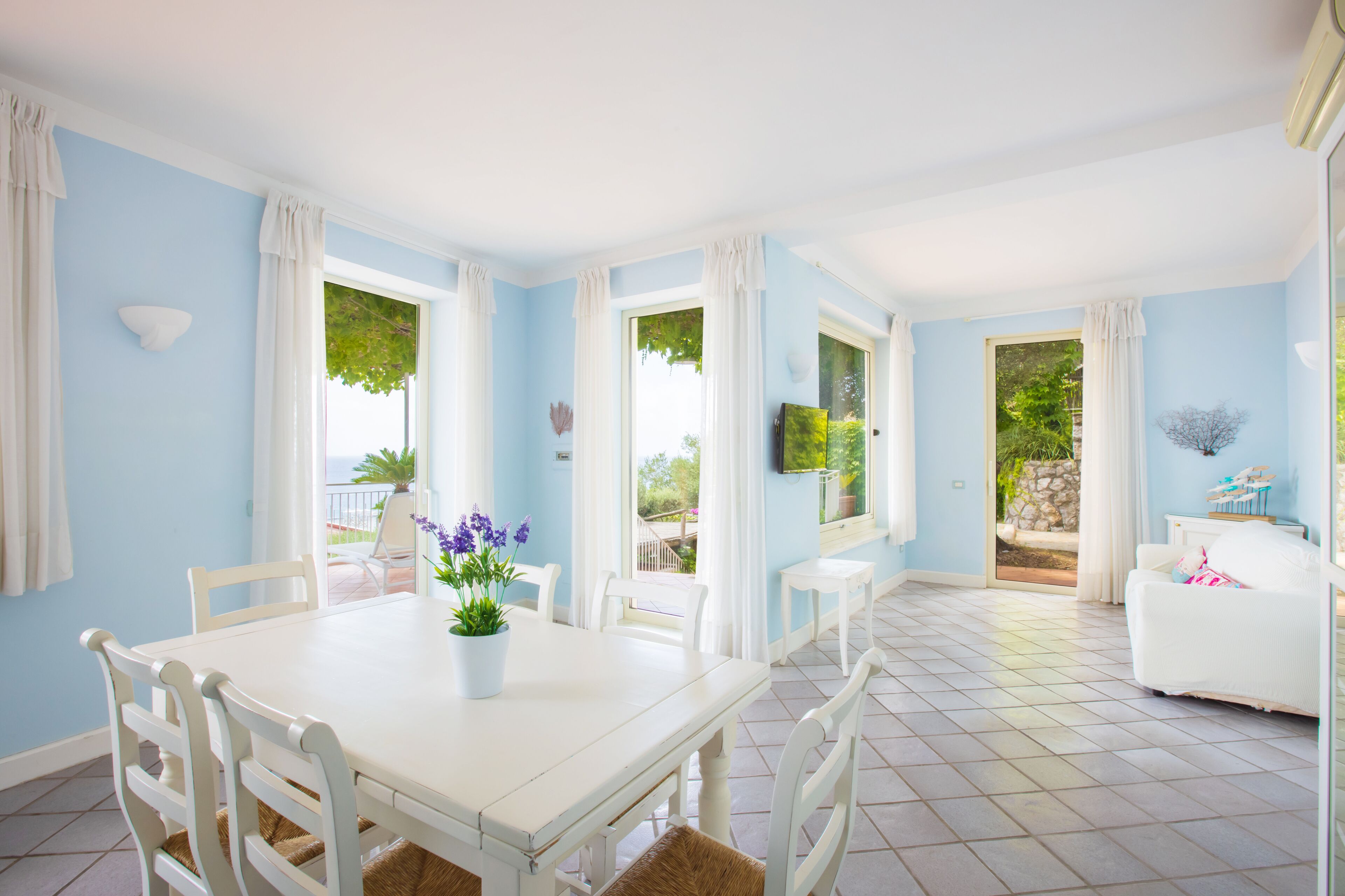 A bright room with a white table, chairs, and a potted plant. There are windows with curtains, a small table, and a white sofa.