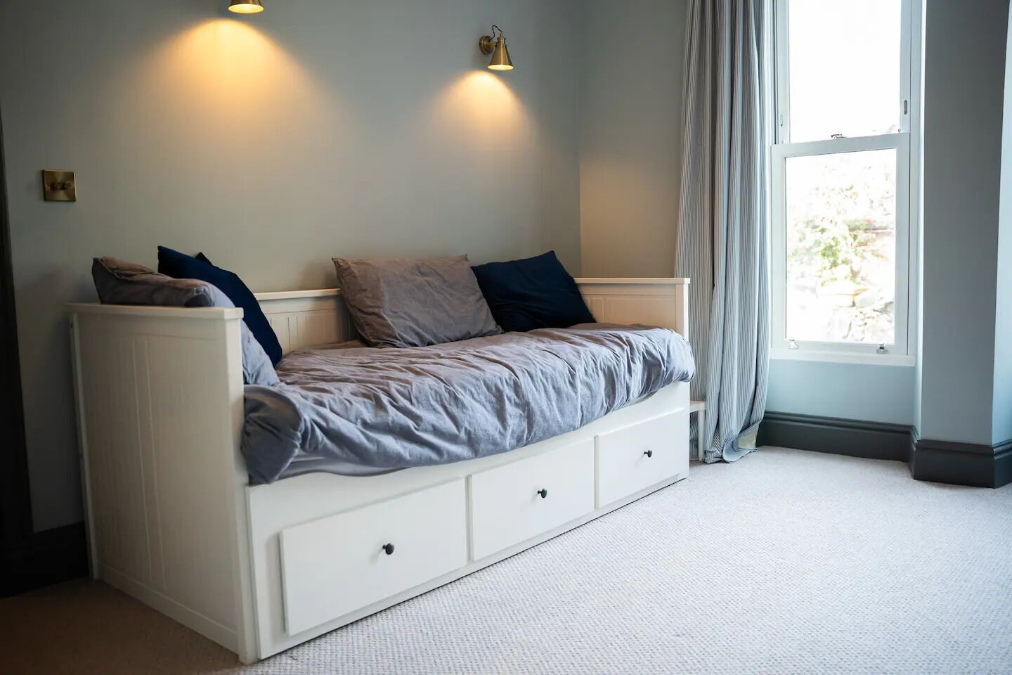 Camera singola con un letto, una panca contenitore integrata, due luci a parete, una finestra con tende e un termosifone.