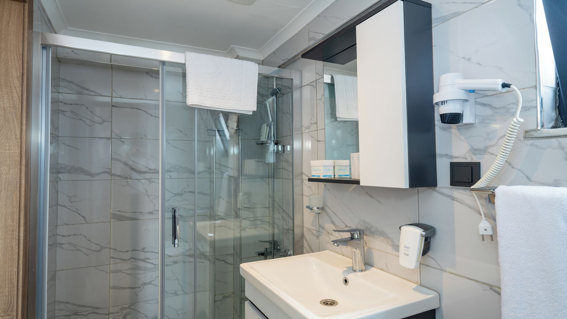 A modern bathroom with a glass shower enclosure, a white sink, and a hairdryer mounted on the wall.