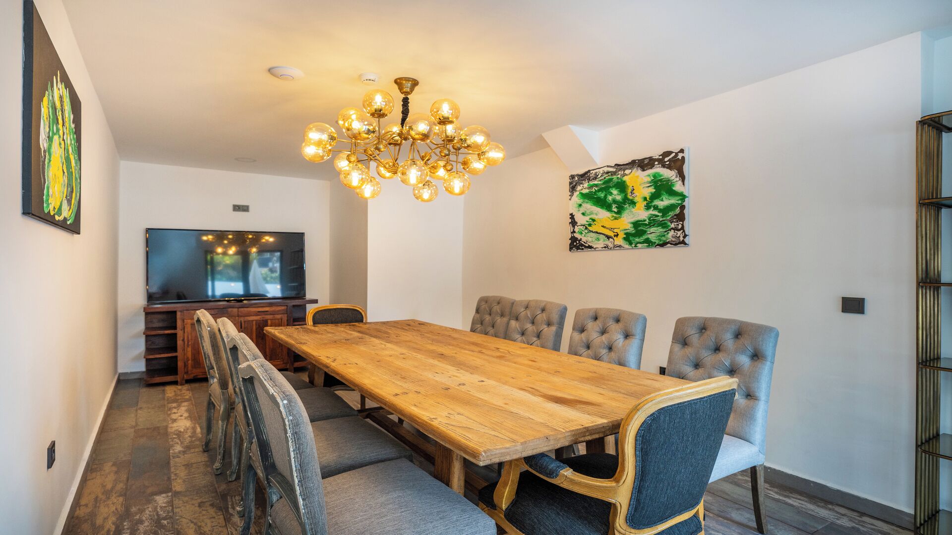 A dining area with a wooden table, six upholstered chairs, a chandelier, a television, and artwork on the walls.