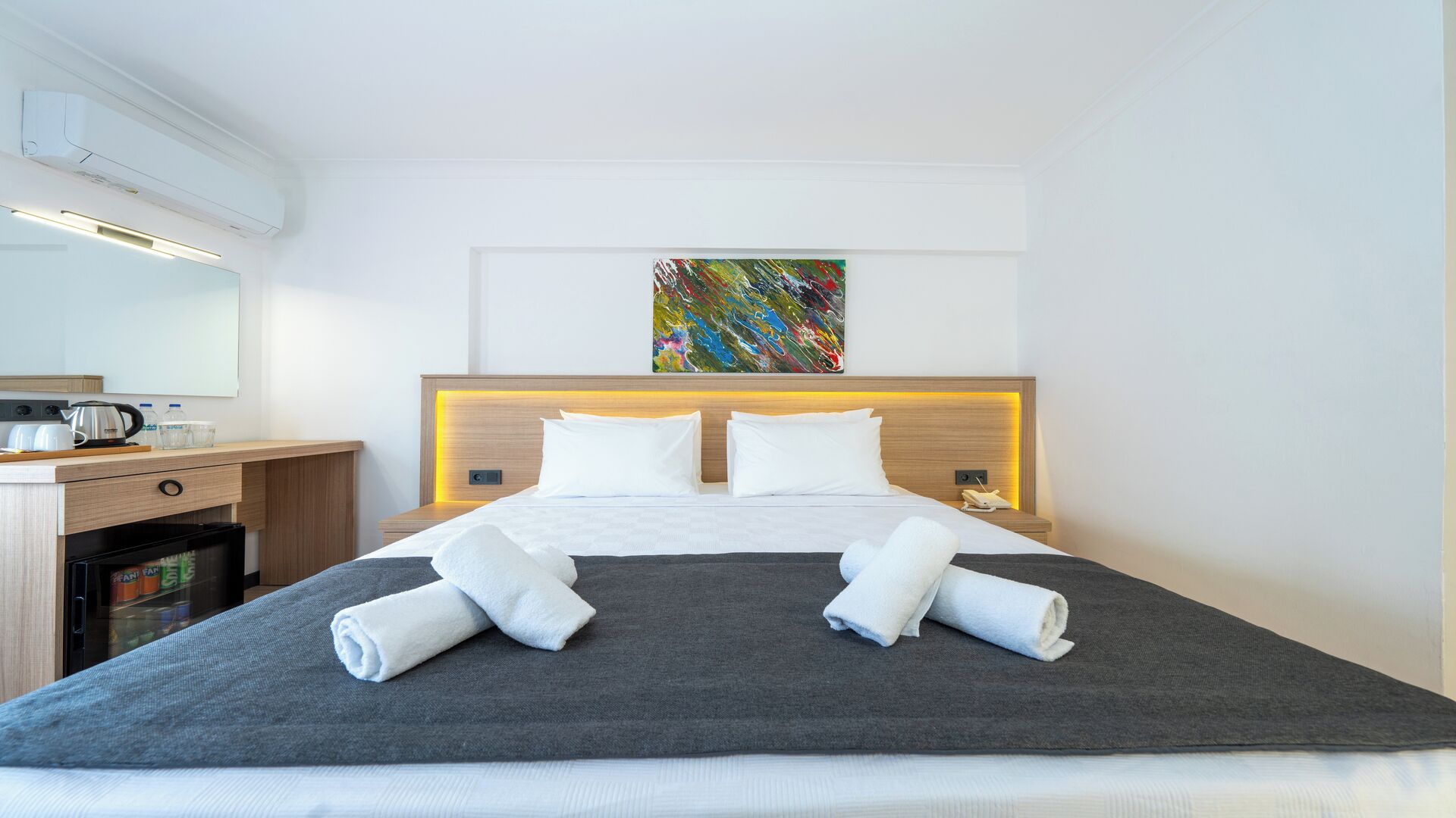 Habitación de hotel con cama, mesita de noche, escritorio pequeño, refrigerador y un colorido cuadro abstracto en la pared.