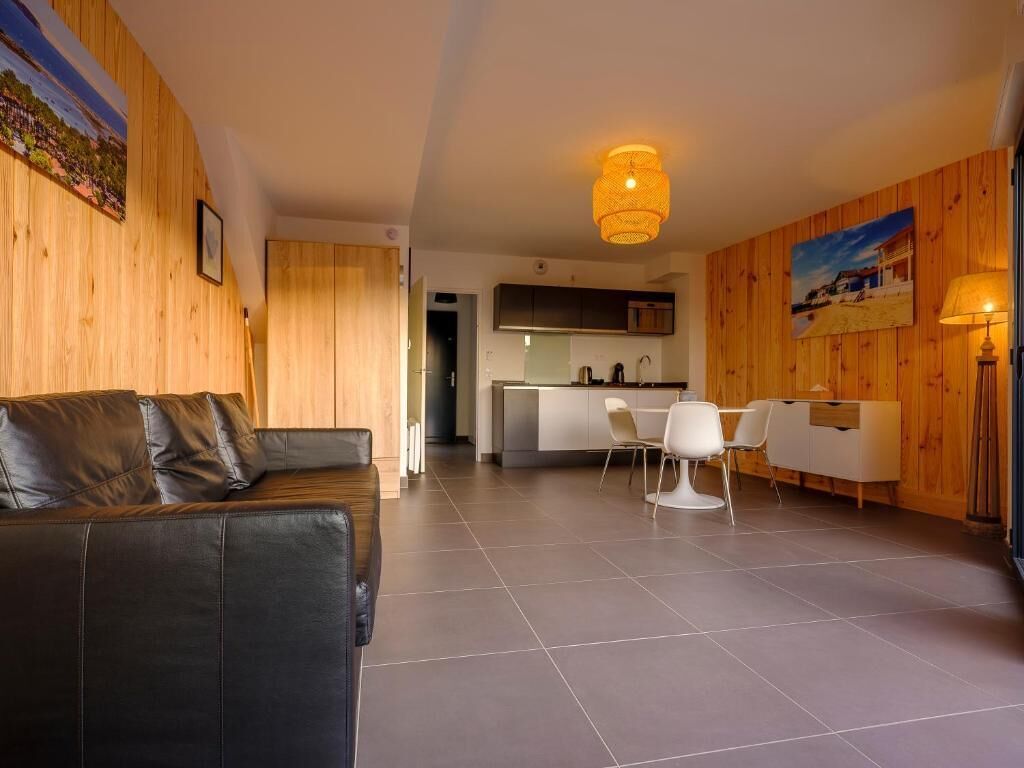 A modern living room with a black leather sofa, a dining area with white chairs, and a kitchen in the background.