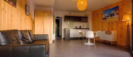 A modern living room with a black leather sofa, a dining area with white chairs, and a kitchen in the background.