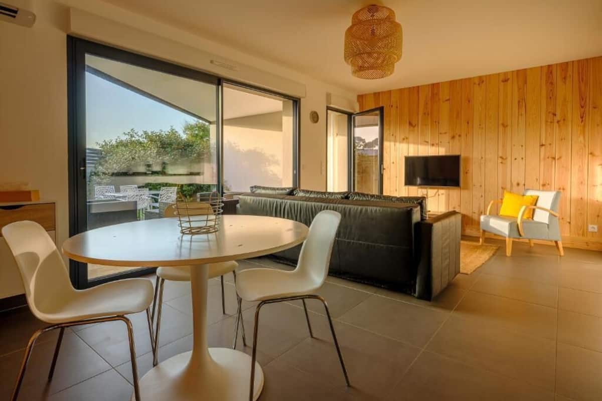 Un salon moderne avec une table ronde, des chaises blanches, un canapé et un téléviseur.