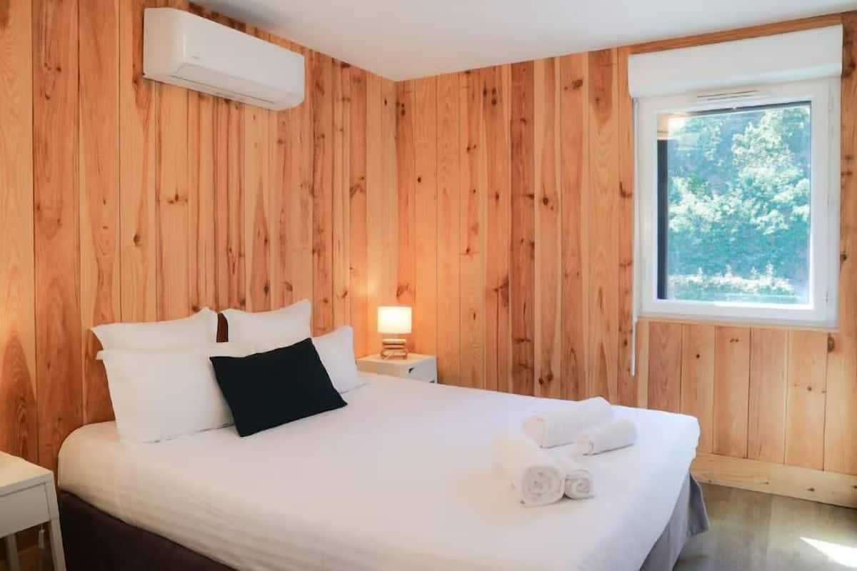 Une chambre à coucher aux murs lambrissés, avec un lit, des oreillers, une table de chevet, une lampe et une fenêtre donnant sur des arbres.