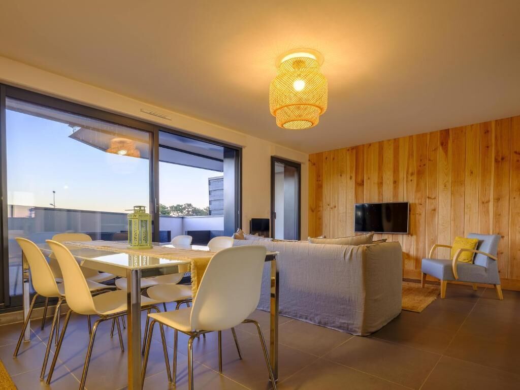 A modern living room with a dining area, wooden panel walls, a flat-screen TV, and a large window offering a view of the city.