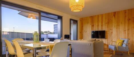 A modern living room with a dining area, wooden panel walls, a flat-screen TV, and a large window offering a view of the city.