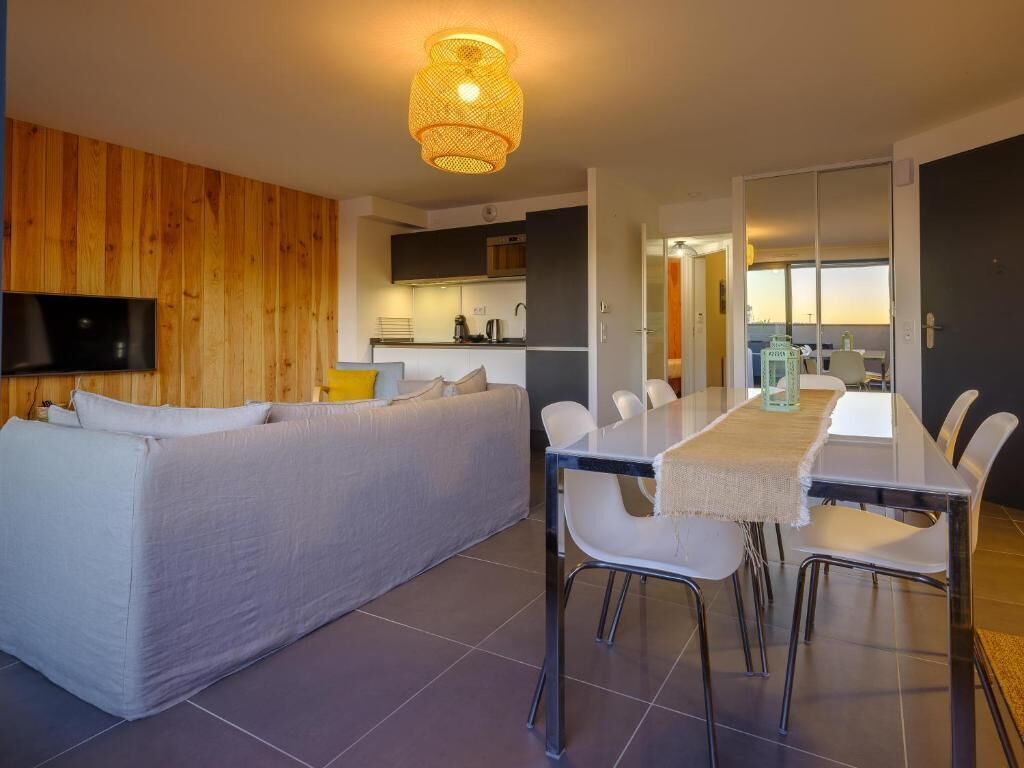 A modern living room with a sofa, dining table, and chairs. There is a kitchen area in the background with a TV mounted on the wall.