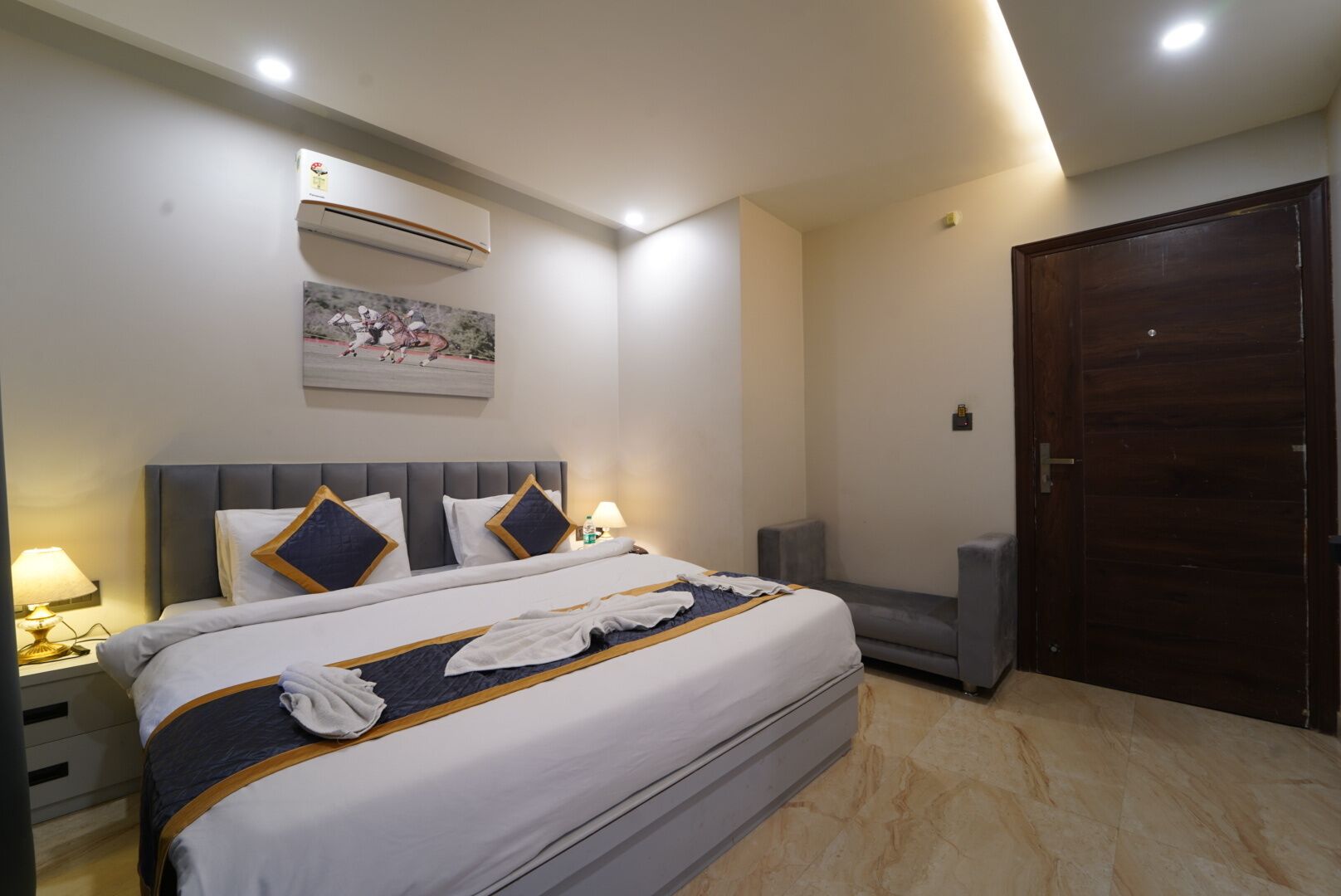 A hotel room with a large bed, a gray upholstered armchair, a wooden door, and an air conditioning unit on the wall.