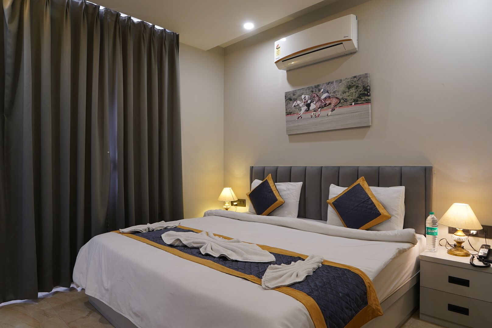A hotel room with a bed, two bedside tables, an air conditioner, a painting on the wall, and a curtain.