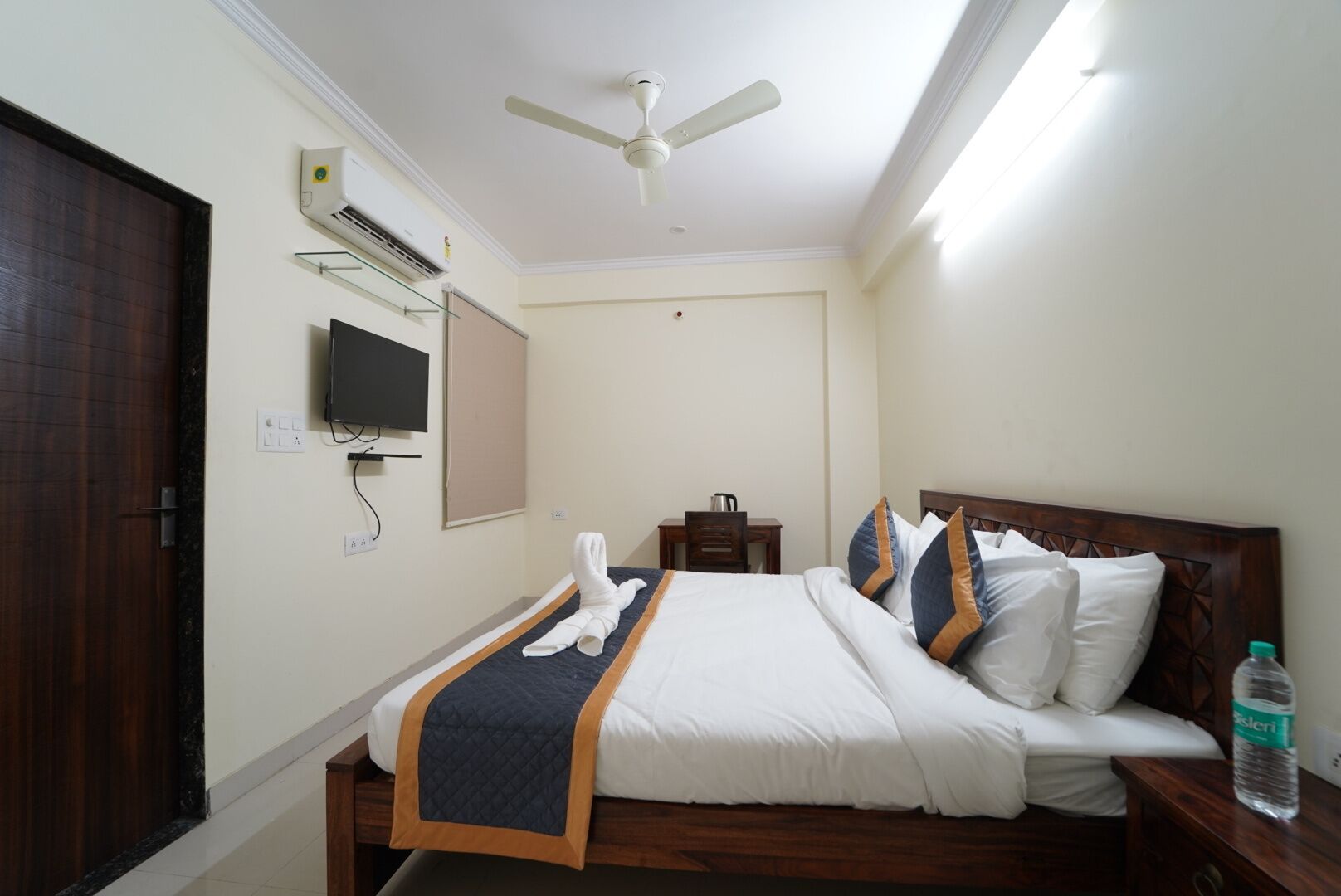 A hotel room with a bed, a desk, a chair, a television, an air conditioning unit, and a water bottle.