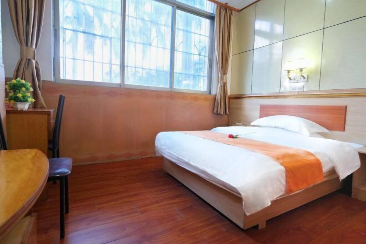 Habitación de hotel con cama, escritorio de madera, una silla, ventana con cortinas y una lámpara fijada a la pared.
