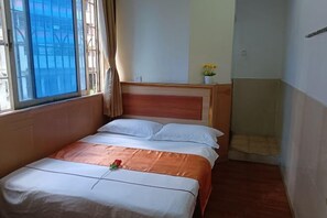 A hotel room with a bed, a window with curtains, a small table with a vase of flowers, and a view of buildings outside. - Xintian Lvye Hotel (Foshan)