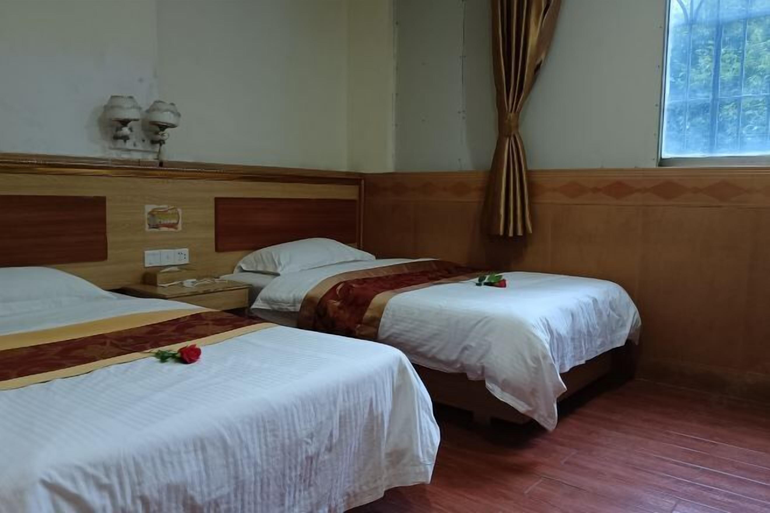 A hotel room with two single beds, wooden headboards, a wooden floor, a window with curtains, and a small table with a lamp.
