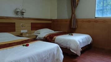 A hotel room with two single beds, wooden headboards, a wooden floor, a window with curtains, and a small table with a lamp.