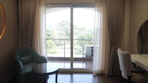 A room with a large sliding glass door leading to a balcony with a view of trees and a building. There is a green armchair, a small round table, and a white chair. - The Wave International Resort, Jamshedpur (Golmuri)