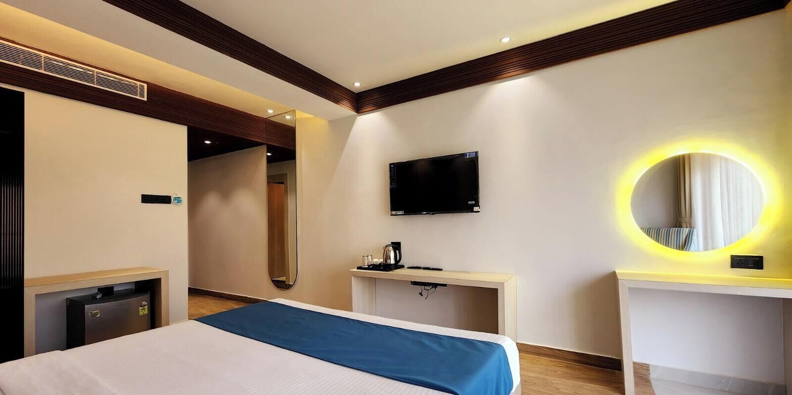 Una habitación de hotel con cama, escritorio, televisor, espejo y una pequeña kitchenette.