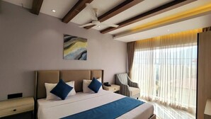 A hotel room with a large bed, a nightstand, a chair, a ceiling fan, and a window with curtains. - The Wave International Resort, Jamshedpur (Golmuri)