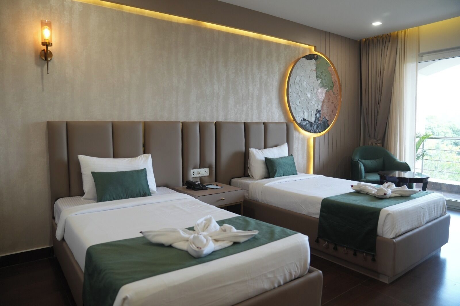 A hotel room with two beds, a round wall clock, a green chair, and a window with curtains.