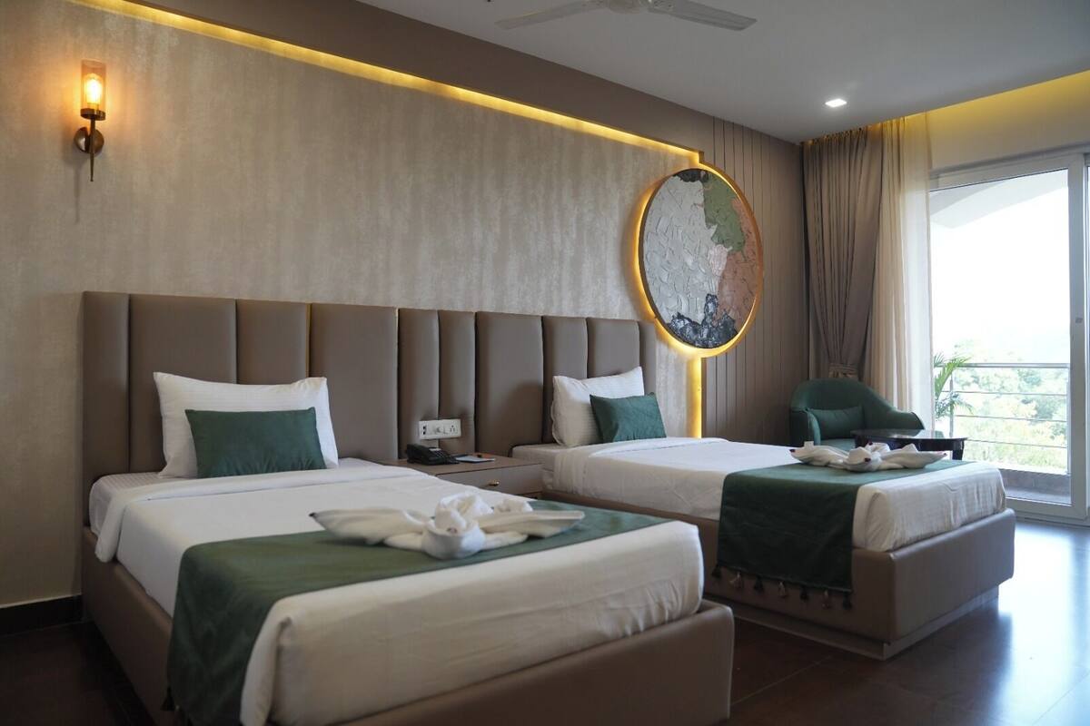 A hotel room with two beds, a large round mirror, a green chair, and a view of greenery outside.