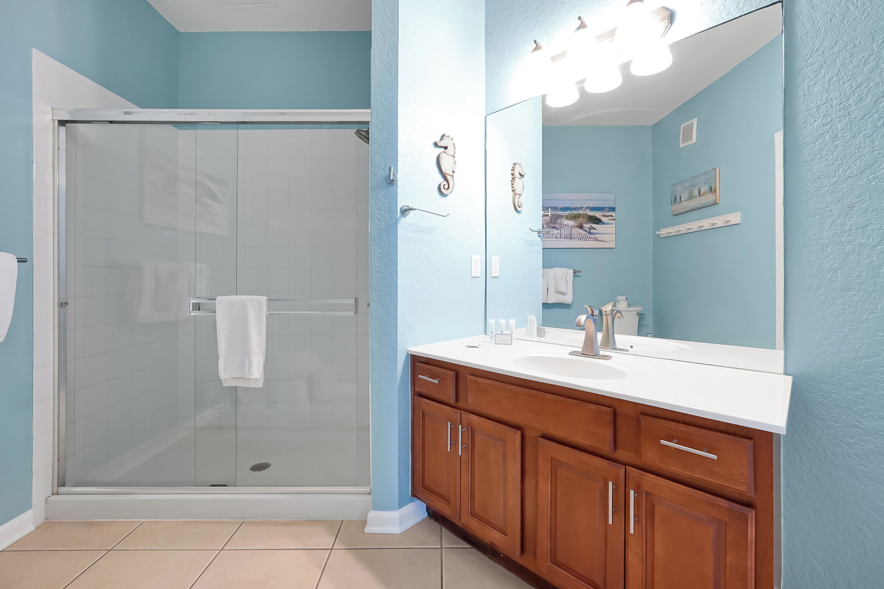 A bathroom with a glass shower enclosure, a white sink with a large mirror, wooden cabinets, and a towel rack.