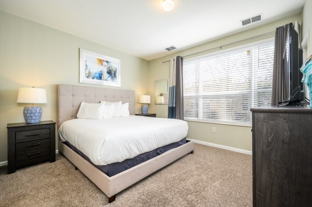 A bedroom with a large bed, two bedside tables, a dresser, a window with blinds, and a ceiling light.