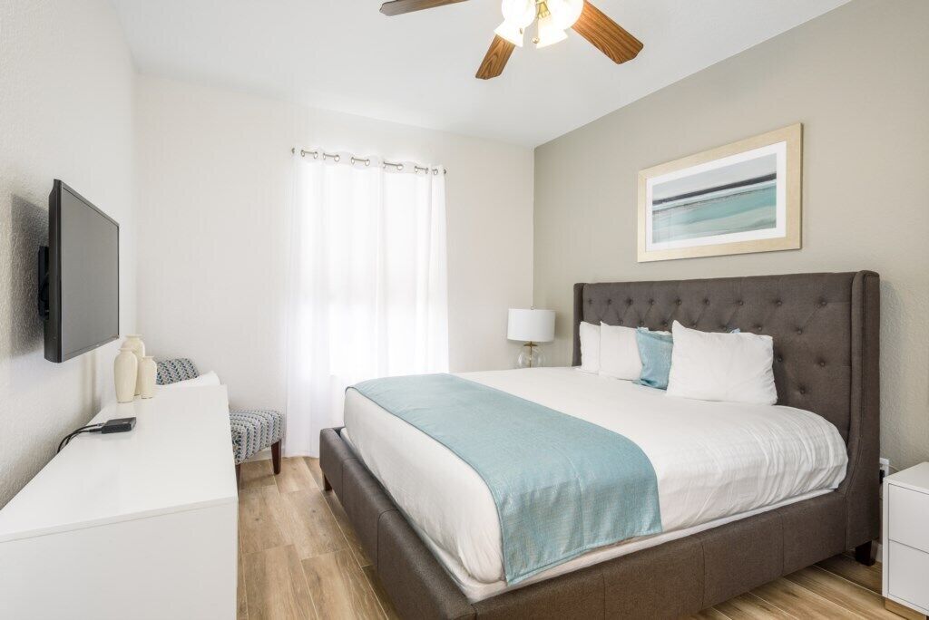 Une chambre moderne avec un grand lit, une table de chevet, un bureau, un ventilateur de plafond et une fenêtre avec des rideaux.