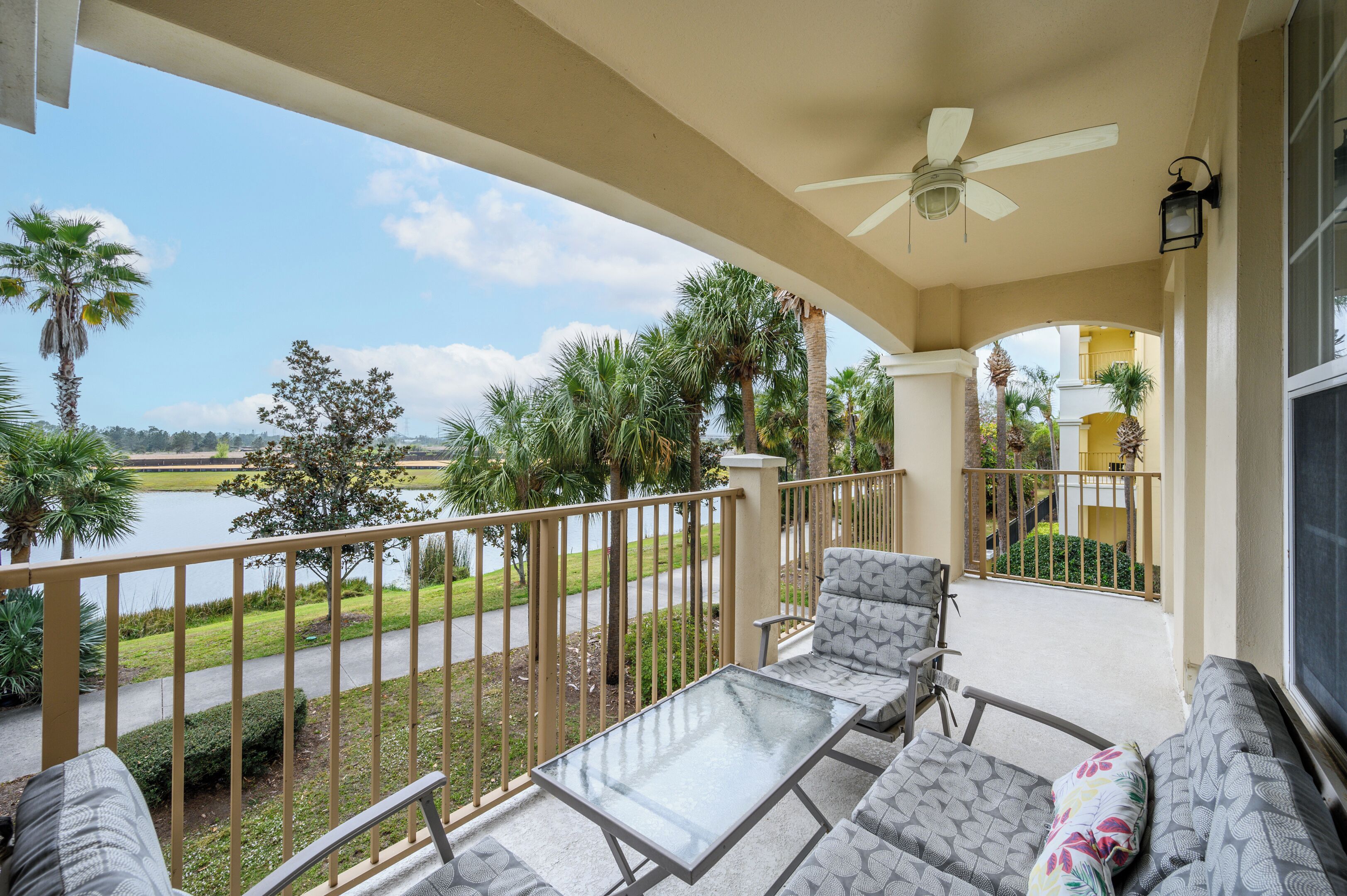 A balcony with a glass table and chairs, overlooking a lake and palm trees.