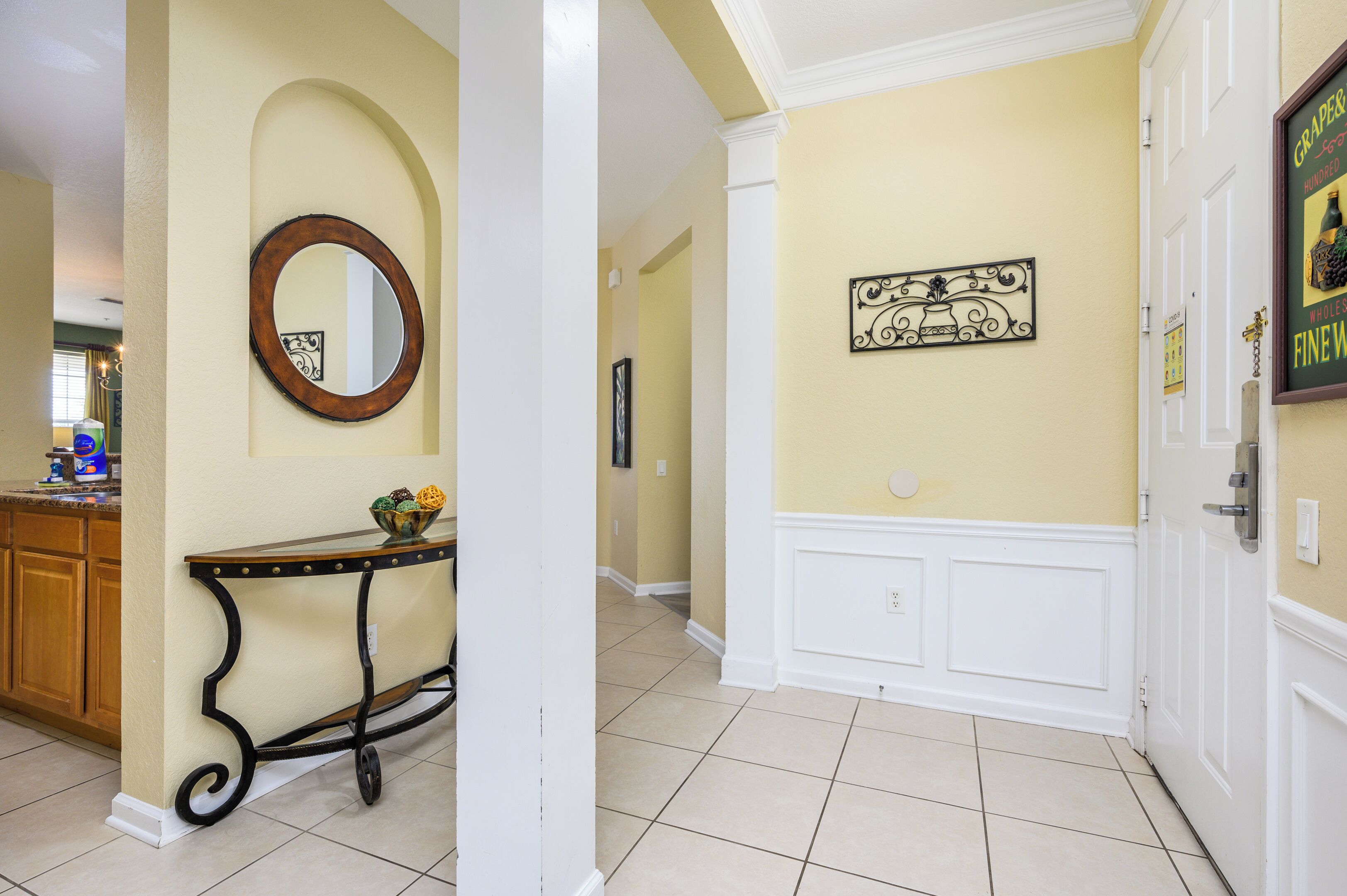 A hallway with a cream-colored wall, a decorative mirror, a small table with a bowl of fruit, and a framed sign.