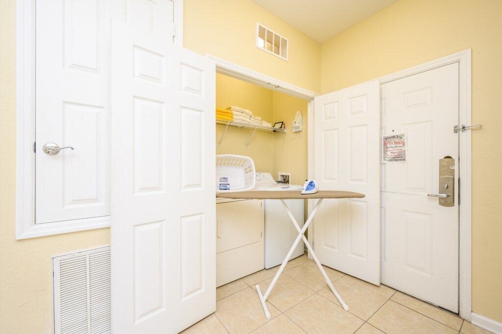 A compact laundry room with a washing machine, dryer, ironing board, and shelves with folded towels.