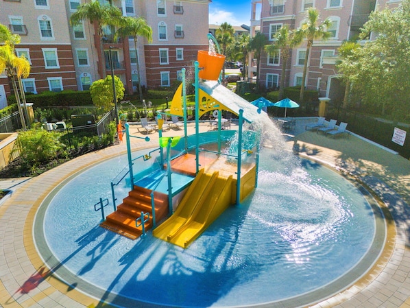 A water play area with slides and a pool, surrounded by a brick building with balconies and palm trees.