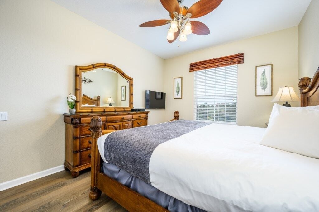 Quarto com cama de madeira, ventilador de teto, cômoda, televisão e janela com persianas.