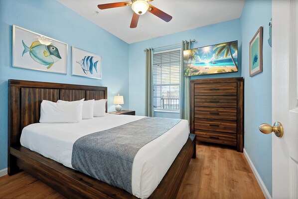 A bedroom with a bed, a nightstand, a ceiling fan, a window with curtains, and wall art.