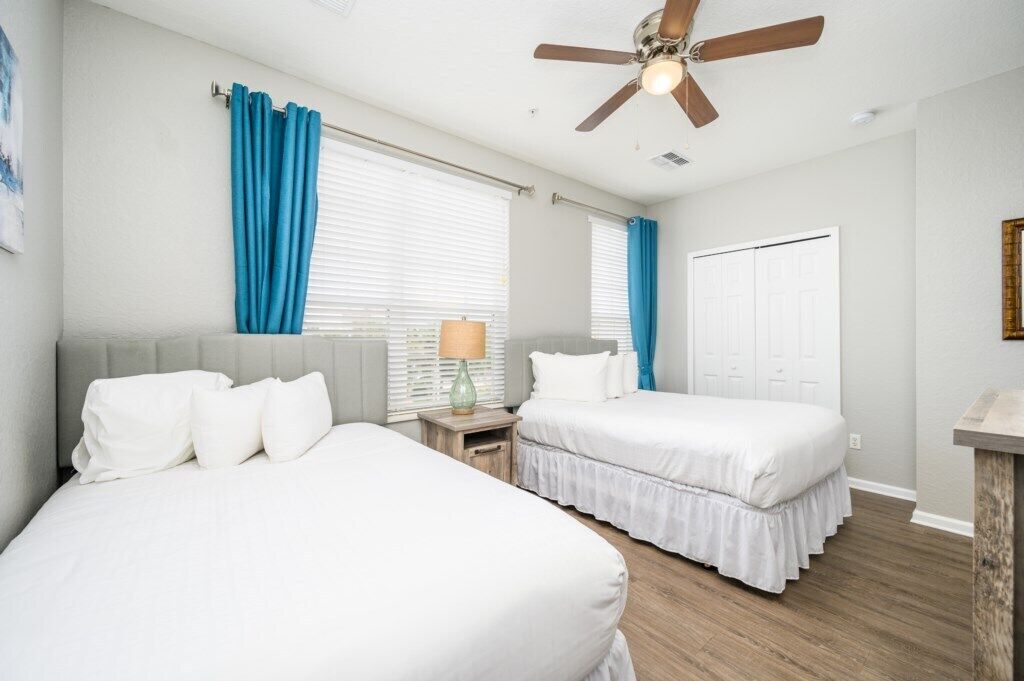 Habitación de hotel con dos camas, ventilador de techo, ventana con cortinas, mesita de noche con lámpara y una puerta.