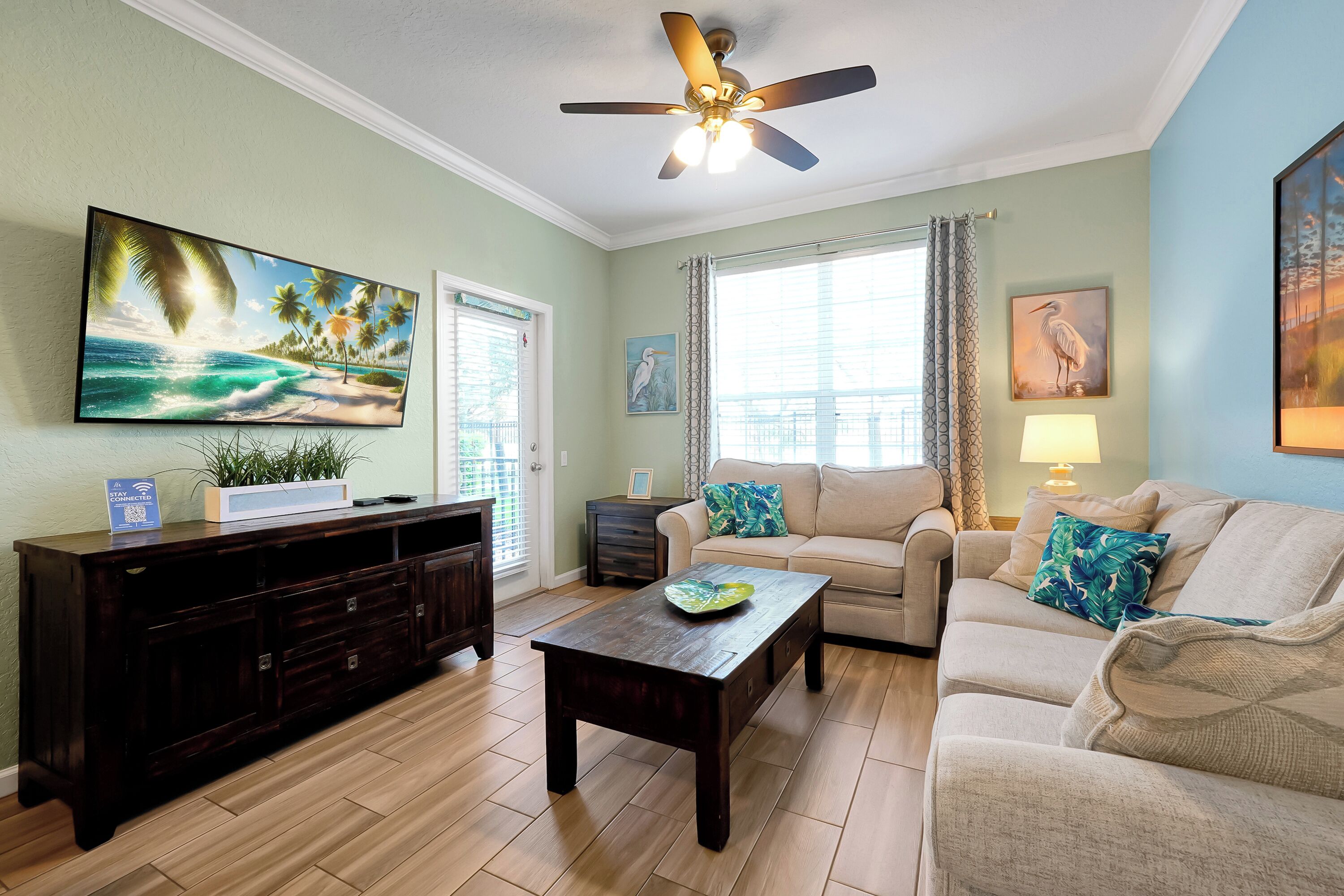 A living room with a dark wooden entertainment center, a beige sofa, a coffee table, a ceiling fan, and a large painting of a beach scene.