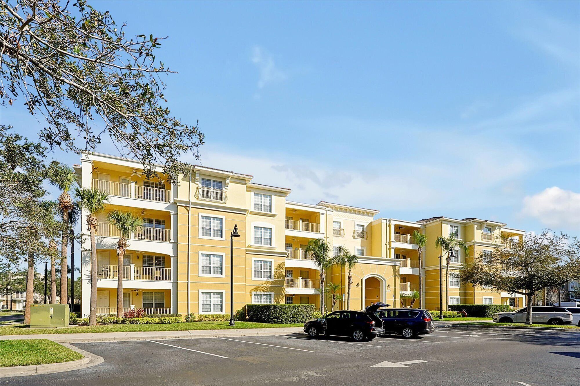 A multi-story yellow building with balconies, a parking lot, and palm trees.