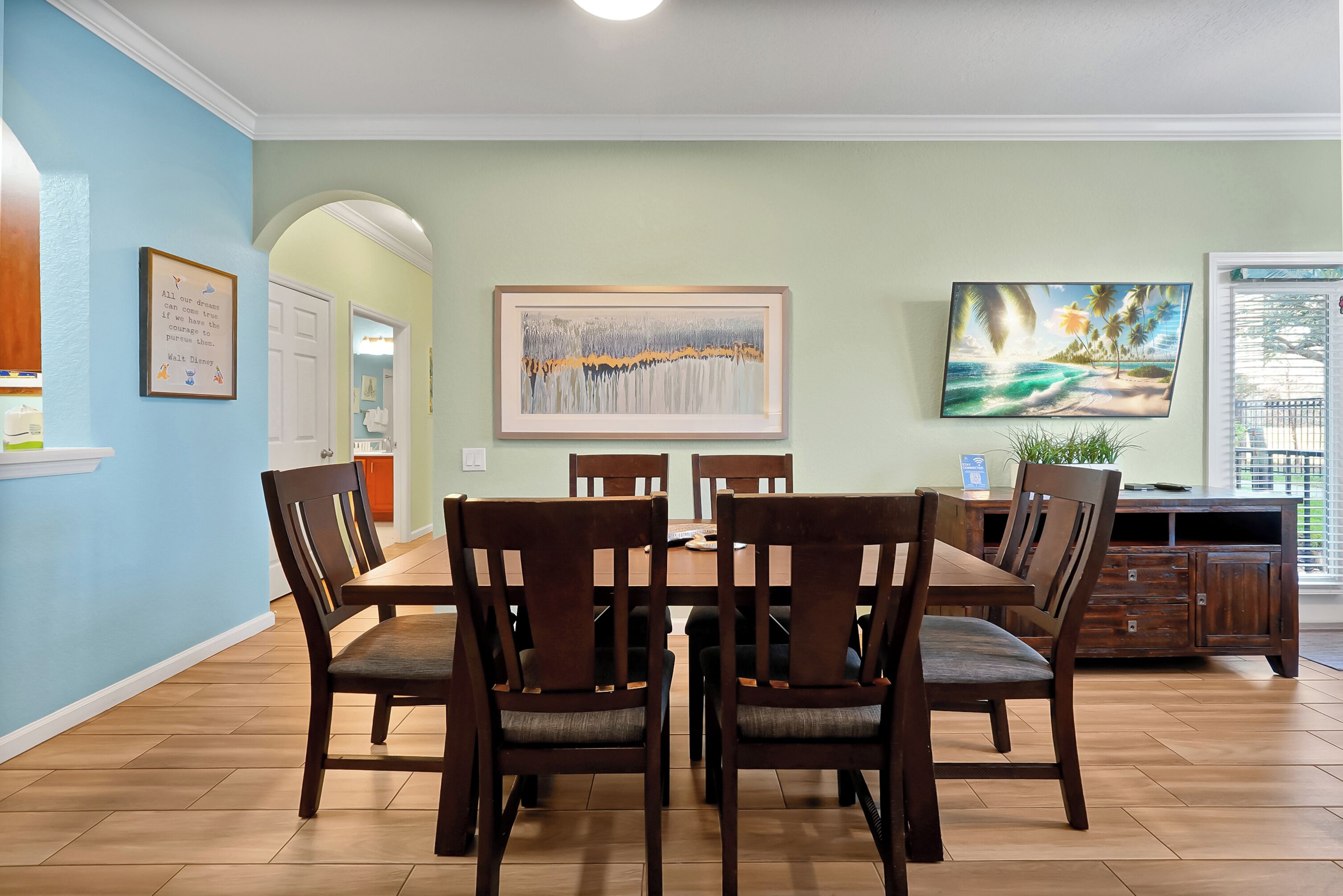 A dining area with a wooden table and chairs, a painting on the wall, and a view of the outdoors through a window.