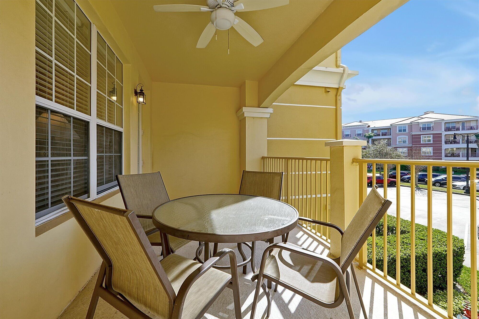 A balcony with a round table and chairs, a ceiling fan, and a view of other buildings and parked cars.