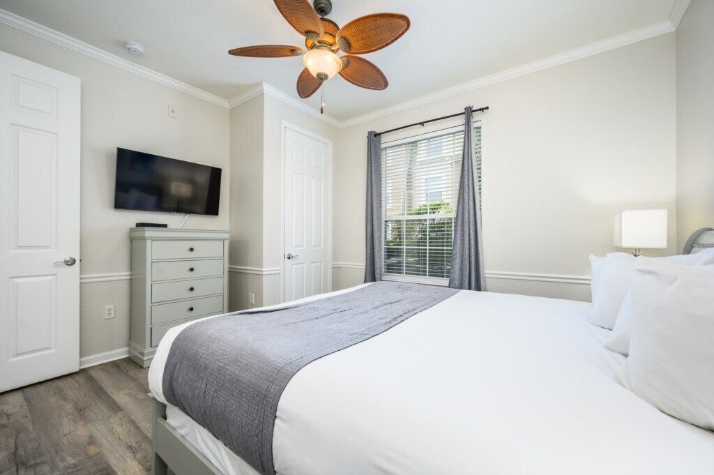 A bedroom with a bed, a ceiling fan, a television, a dresser, and a window with blinds.