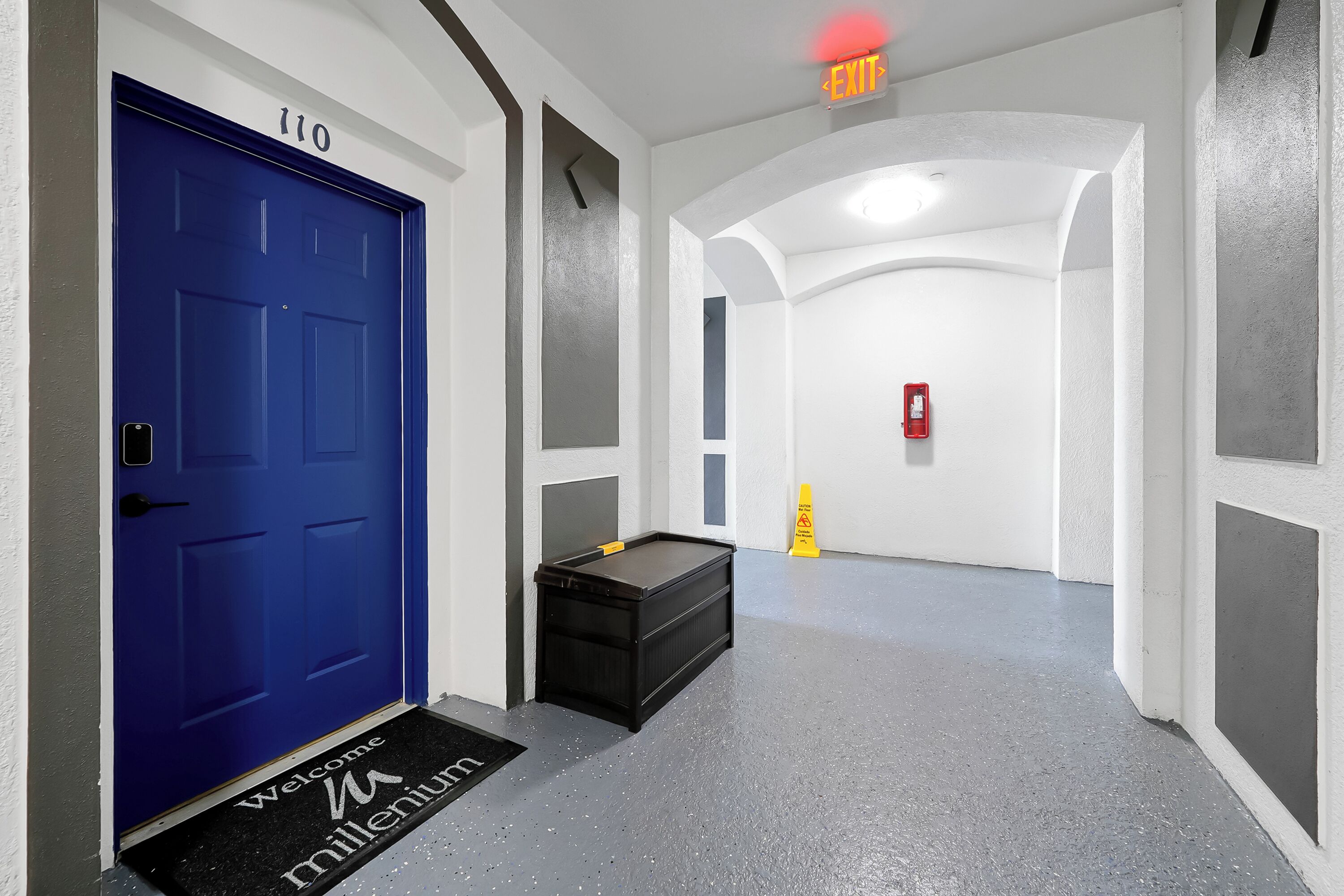 A hallway with a blue door labeled '110', a black bench, a yellow caution sign, and a fire extinguisher on the wall.