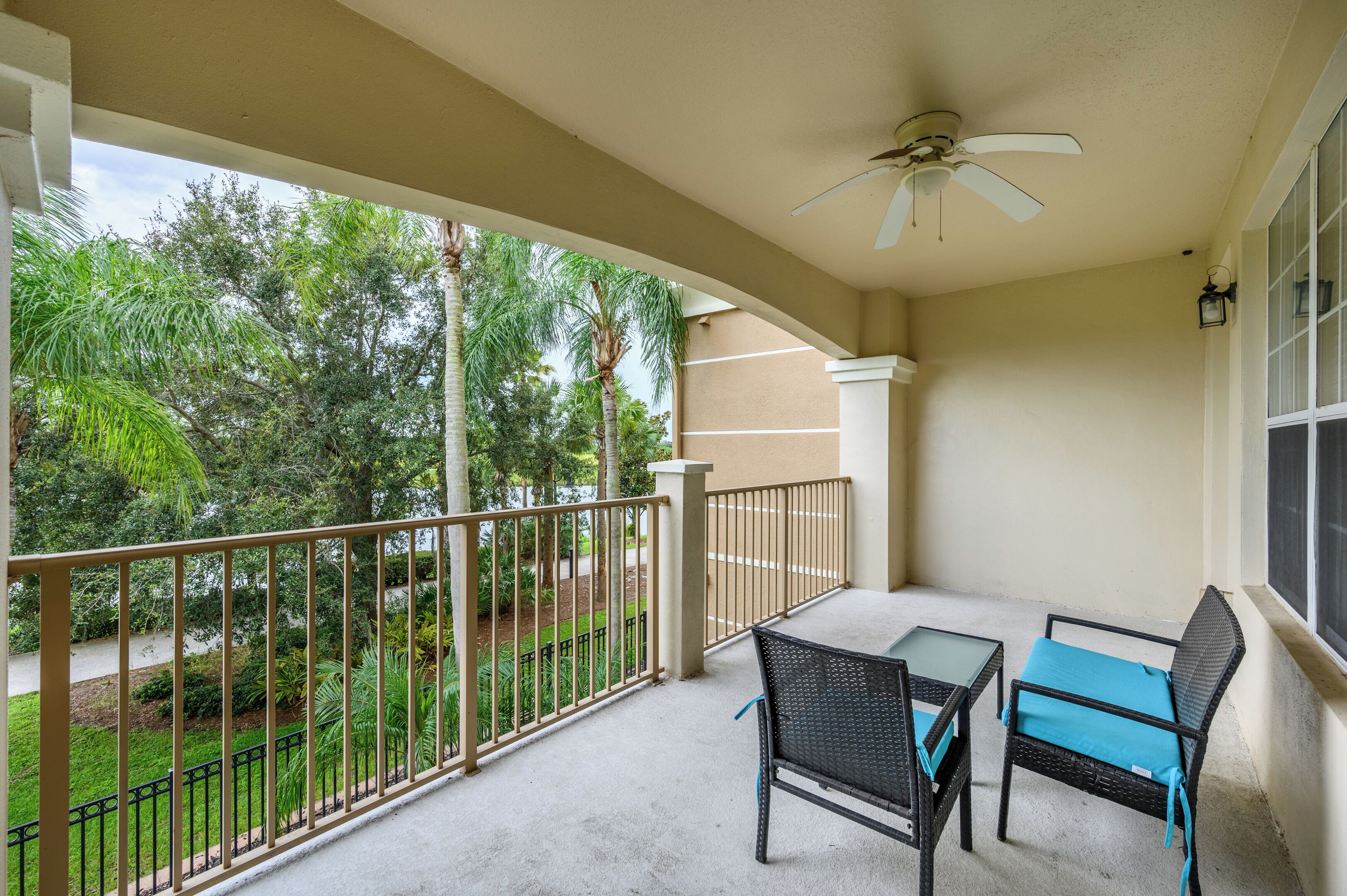 A balcony with a table and two chairs, a ceiling fan, and a view of a landscaped garden with trees.
