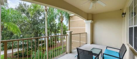 A balcony with a table and two chairs, a ceiling fan, and a view of a landscaped garden with trees.