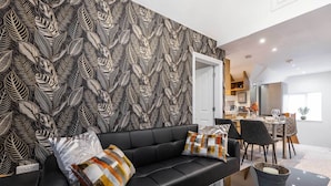 Een moderne woonkamer met een zwarte leren bank, behang met een patroon, en een eethoek op de achtergrond.